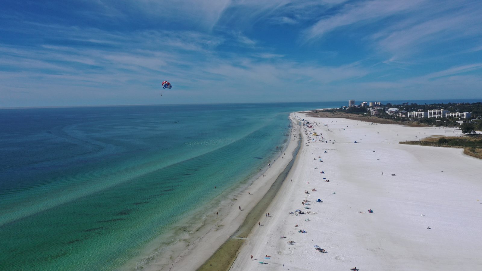 Aerial view north along Siesta Beach, a parasail over the Gulf and the shoreline curving toward Siesta Key Village in the distance
